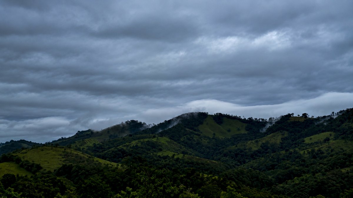 Lush green mountain ridges shrouded in mist beneath a vast, dramatic sky filled with dark storm clouds.